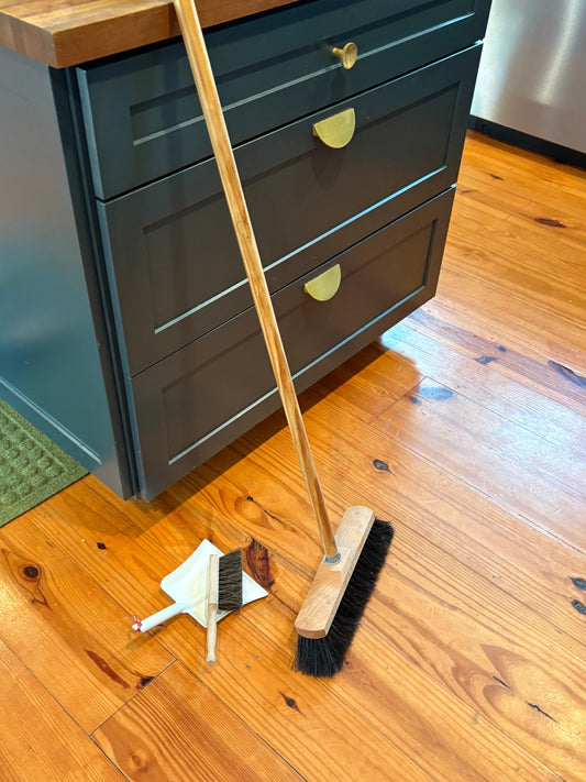 Horsehair and wooden broom on wooden floor next to a small metal dustpan and wooden brush