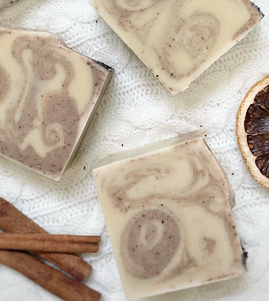 Three swirled soap bars on a textured surface with cinnamon sticks and a dried fruit.